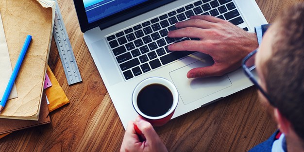 En man som holder én hånd på tastaturet på laptopen sin, og den andre hånden holder en kaffekopp. Foto