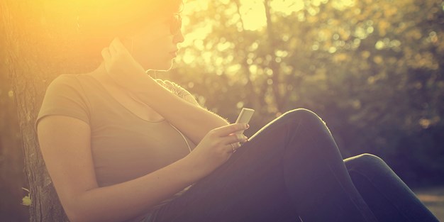 Ung kvinna som sitter i solen med sin mobiltelefon. Fotografi